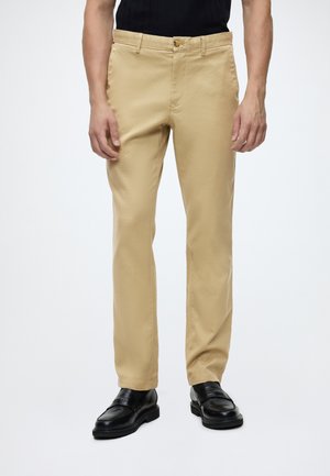 Mand iført beige, lige ben bukser, sort skjorte og sorte læder loafers, stående mod en hvid baggrund.