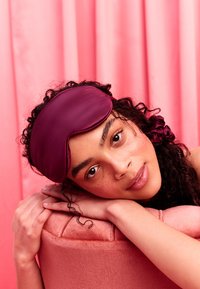 Młoda kobieta z kręconymi włosami odpoczywająca na różowej poduszce, nosząca fioletową maskę do spania przesuniętą na czoło, w tle różowa zasłona.