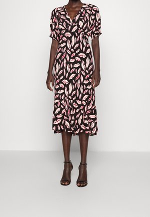 Kobieta w czarnej sukience midi z różowo-czerwonymi nadrukami w kształcie ust, z bufiastymi krótkimi rękawami i czarnymi szpilkami z paskiem na kostce, stojąca na szarej podłodze.