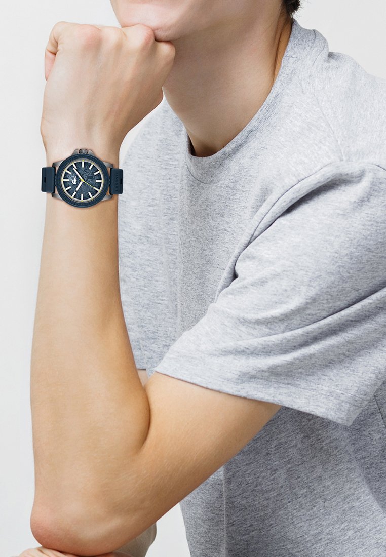 Personne en t-shirt gris reposant son menton sur le poing, portant une montre bleu foncé avec un cadran texturé et une trotteuse jaune au poignet gauche.