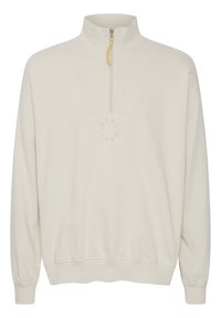 Sudadera beige con alto cuello y media cremallera, con una textura suave y un logo circular en el centro. Puños y dobladillo acanalados.