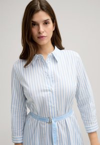 Abito camicia a righe blu chiaro e bianche con bottoni frontali, colletto, maniche 3/4 e vita con cintura caratterizzata da una fibbia circolare in oro.