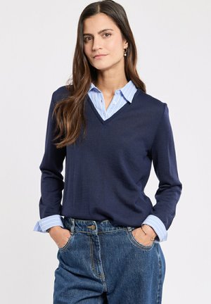 Maglione a v blu navy realizzato in tessuto morbido, abbinato a una camicia a righe blu chiaro con bottoni al colletto e ai polsini, indossato con jeans di denim.