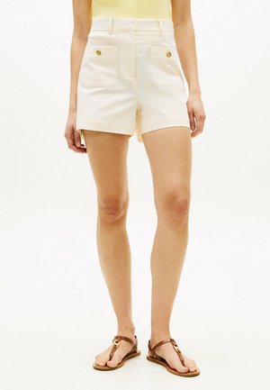 Frau trägt cremefarbene High-Waist-Shorts mit vorne geknöpften Taschen und braunen flachen Ledersandalen, steht vor einem einfarbigen Hintergrund.