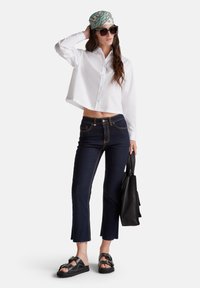 Camicia bianca corta con bottoni, jeans a zampa d'elefante in denim scuro con cuciture a contrasto, sandali neri spessi, foulard decorato e borsa nera.