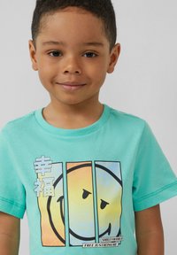 Turquoise T-shirt met een grafisch ontwerp van een glimlachend gezicht, tekst in het wit en accenten van geel en blauw. Korte mouwen en ronde hals.