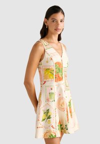 En kvinne poserer i en ermeløs, mønstret kjole med livfulle frukt- og blomstermotiver, mot en lys grå bakgrunn.