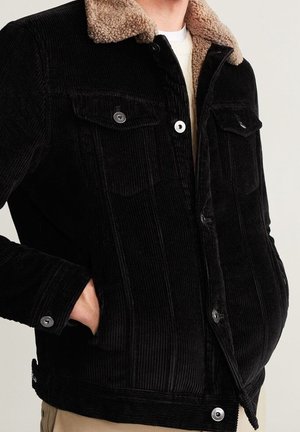 Czarna sztruksowa kurtka z beżowym kołnierzem sherpa, noszona na białej koszuli, ręce schowane w bocznych kieszeniach, pokazana od klatki piersiowej do bioder.