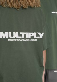 Grøn bomulds-T-shirt med stor hvid tekst på ryggen, der lyder "MULTIPLY" og "MULTIPLY APPAREL CLUB" i fed skrift.