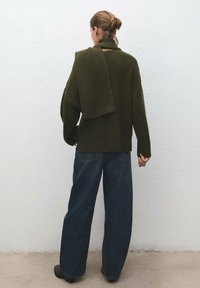 Maglione oversize verde scuro lavorato a maglia con una sciarpa abbinata drappeggiata sulla spalla, abbinato a jeans larghi in denim blu scuro.