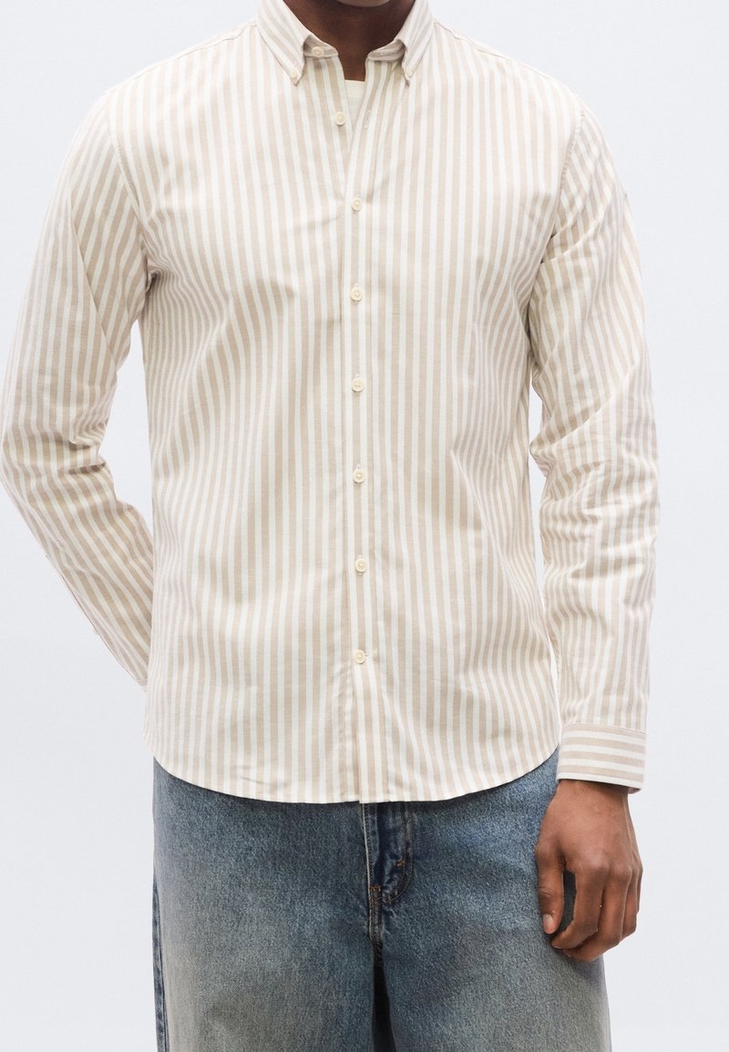 Homme portant une chemise beige et blanche à rayures verticales avec boutons et manches longues, et un jean bleu clair sur fond uni.