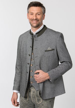 Mann lächelnd in traditioneller grauer Jacke mit Holzknöpfen, weißem Hemd, Weste und bestickten Lederhosen vor einfarbigem Hintergrund.