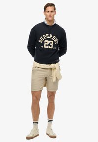 Sudadera marina con letras crema "SUPERDRY 23", pantalones cortos beige, suéter crema atado a la cintura, zapatos crema y calcetines a rayas azul claro.