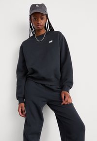 Ensemble sweat-shirt et pantalon noir, avec un col rond, des poignets côtelés et un logo blanc. Associé à une casquette grise et un collier en chaîne argenté.