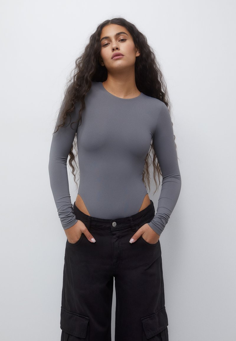 Donna con capelli ricci lunghi che indossa un body grigio a maniche lunghe aderente e pantaloni cargo neri larghi con le mani in tasca.