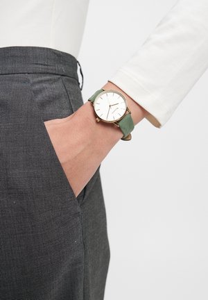 Grüne Lederarmbanduhr mit rundem goldenen Gehäuse und weißem Ziffernblatt; am Handgelenk getragen, kombiniert mit grauen strukturierten Hosen.