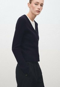 Cárdigan de punto en color navy con un patrón texturizado, cuello en V y cierre de botón. Combinado con una blusa blanca y pantalones negros de corte ajustado.