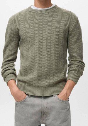 Mann trägt einen hellolivgrünen, gerippten Strickpullover mit Chevronmuster und hellgraue Jeans, Hände in den Taschen.