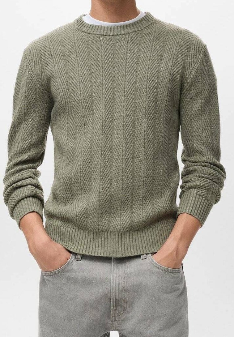 Homme portant un pull en maille côtelée vert olive clair avec un motif chevron et un jean gris clair, mains dans les poches.