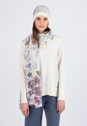 Bloemenpatroon sjaal en muts in roomkleur, met paarse, roze en gele accenten. Zachte gebreide textuur, ontworpen voor warmte. Model draagt een casual sweater en broek.