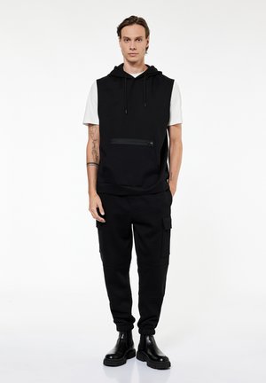 Mężczyzna stoi, ubrany w czarną bezrękawnikową bluzę z kapturem na białą koszulkę z krótkim rękawem, czarne spodnie typu cargo i czarne buty, na białym tle.