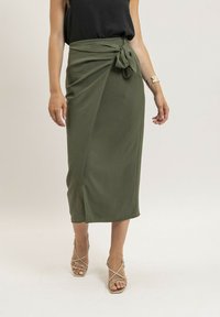 Jupe portefeuille vert olive en tissu doux, dotée d'un détail de nœud à la taille et d'une longueur midi, associée à des sandales dorées à lanières.