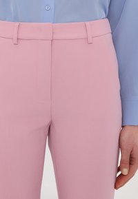 Femme portant un pantalon rose clair et une chemise bleu clair, avec des passants de ceinture visibles et un poignet de chemise partiellement boutonné.