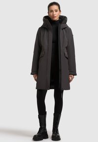 Parka nera impermeabile con cappuccio staccabile, due tasche frontali, maniche lunghe e un design elegante abbinata a leggings neri e stivali.