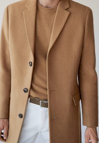 Abrigo de lana de camello con solapas de muesca, dos botones y bolsillos laterales. Superpuesto sobre un suéter beige, combinado con pantalones blancos y un cinturón verde.