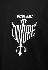 Μαύρο ύφασμα με λευκό στυλιζαρισμένο κείμενο "Versace Jeans Couture" και ένα βέλος με τρεις αιχμές που σχηματίζουν το λογότυπο.