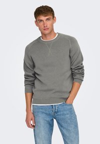 Pull en maille gris avec un col rond, des manches longues et des poignets côtelés. Porté sur un sous-vêtement blanc, associé à un jean en denim bleu.