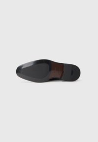 Suela de un zapato de vestir negro con acabado pulido, sección media marrón marcada con la marca "BOSS" y tacón de goma, fotografiada sobre un fondo blanco.