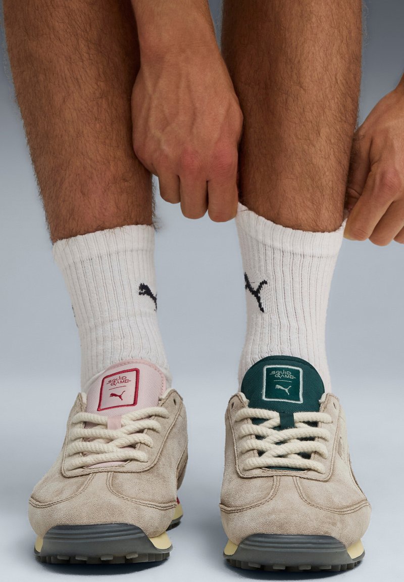 Le sneaker in suede beige presentano dettagli rosa e verdi sui talloni, lacci spessi e una suola in gomma testurizzata. Sono indossati calzini bianchi a coste.