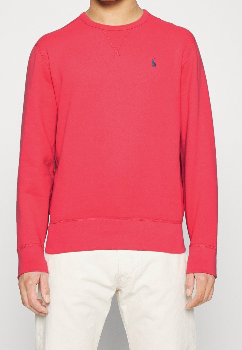 Homme portant un sweat-shirt rouge uni à manches longues avec un petit logo bleu sur la poitrine et un pantalon de couleur claire.