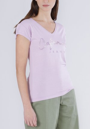 Mujer con camiseta de manga corta color lila con cuello en V y texto "Cafenoir Jeans" y pantalones verde claro, sobre un fondo liso.