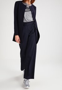 Marinblazer över grå Nike-t-shirt, parat med randiga vida byxor och silverfärgade sneakers, vilket visar en lager-på-lager avslappnad stil.