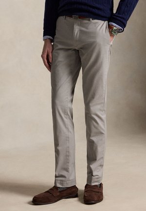Homme portant un pantalon beige, un pull bleu marine, des mocassins en daim marron et une montre-bracelet, debout avec une main dans la poche.