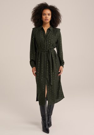 Robe chemise verte à motifs avec col, manches longues, ceinture à la taille et fentes sur les côtés, associée à des bottes noires jusqu'au genou.