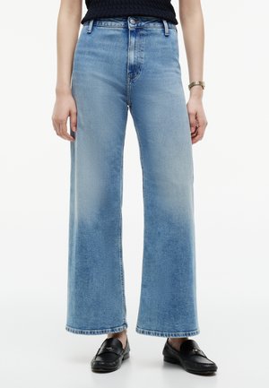Kvinde iført lyseblå jeans med høj talje og brede ben, sorte loafers, sort ærmeløs top og guld armbåndsur mod en hvid baggrund.