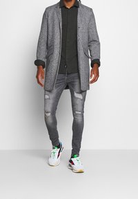 Grå uldfrakke over en mørk skjorte med knapper, parret med slidte grå jeans og hvide sneakers med farverige accenter.
