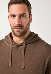 Bruine hoodie met een textuurstof, voorzien van een ribbelontwerp en trekkoorden. De kleur is een rijke, warme bruine tint.