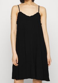 Černé šaty bez rukávů s volným střihem, s volánkovým výstřihem a tenkými ramínky. Materiál vypadá lehký a strukturovaný.