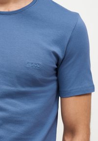 Homme portant un t-shirt bleu ajusté à manches courtes avec "BOSS" brodé sur la poitrine gauche.
