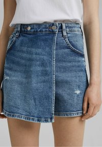 Denimrock in mittlerem Blau mit hoher Taille, zwei Vordertaschen, ausgewaschenen Details und kontrastierenden Nähten entlang des Saums.