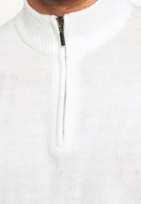 Weißer Strickpullover mit hohem Kragen und halbem Reißverschluss. Die Textur ist weich und hat ein dezentes gewebtes Muster.