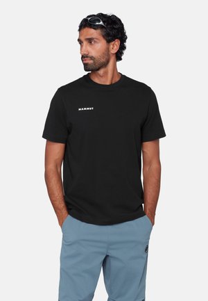 Homme aux cheveux courts et bouclés, de couleur foncée, portant un T-shirt noir Mammut et un short bleu clair, les mains dans les poches, regardant vers sa gauche.