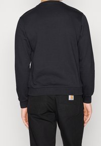 Felpa nera con maniche lunghe e collo tondo, caratterizzata da polsini e orlo ribattuti. La vista dal retro mostra il logo Carhartt sulla vita.