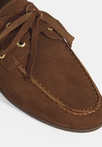 Braune Wildlederslipper mit runder Zehenform, ausgestattet mit goldenen Ösen, passenden Schnürsenkeln und kontrastierenden Nähten entlang des Randes.
