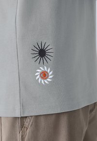 Pilkas marškinėlis su juodu saule ir baltais gėlių siuvinėjimais su oranžinėmis detalėmis, pagamintas iš medvilnės su minkšta tekstūra.