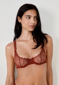Reggiseno in pizzo di colore ruggine, caratterizzato da motivi floreali trasparenti, bordi a scallop e spalline regolabili, progettato con supporto ferretto.